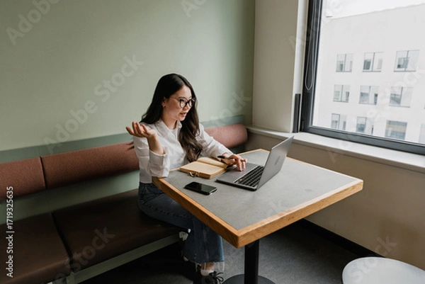 Fototapeta A female worker scrolls on a laptop and talks while sitting at the table