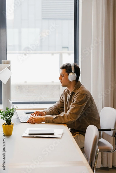 Fototapeta Male worker listens in headphones and looks at laptop while sitting at the table