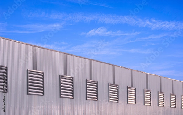 Fototapeta Large industrial factory production building with row of ventilation louvers on gray aluminium corrugated steel wall against blue sky background, Side view with copy space