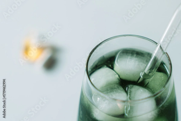 Fototapeta Tight crop of the chlorophyll iced glass with a glass straw, vitamins out of focus in the distance. Clean minimal look. Horizontal.