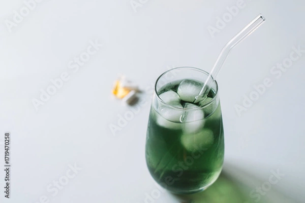 Fototapeta Angled top view of an iced chlorophyll drink with a glass straw, vitamins blurred in the background. Horizontal with copy space.