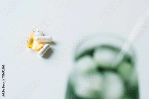 Fototapeta Vitamins in sharp focus, capsules, tablet, softgel, on a light surface, green drink blurred in the background. Horizontal with copy space.