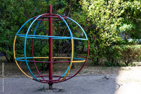 Fototapeta A vibrant, colorful spinning playground orb stands in a park, its bright red, blue, and yellow rings contrasting with the green bushes and dappled sunlight.

