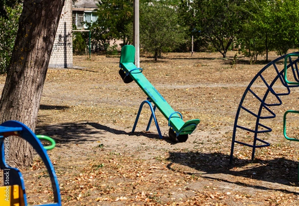 Fototapeta A lonely seesaw sits on a playground in a sunlit park, surrounded by dry grass and fallen leaves. The image captures a quiet, nostalgic moment of childhood and summer.

