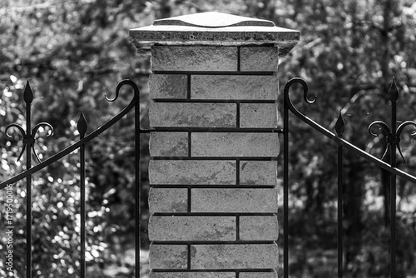 Fototapeta A detailed, close-up, monochrome shot of a brick pillar and an ornate wrought iron fence against a blurred background of trees, highlighting textures.

