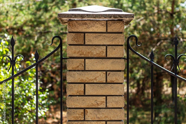 Fototapeta A detailed, close-up shot of a brick pillar with a decorative wrought iron fence on a sunny day, with soft-focus green foliage in the background.

