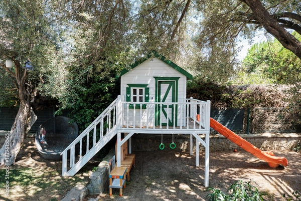 Fototapeta White and green playhouse with slide in a garden under olive trees