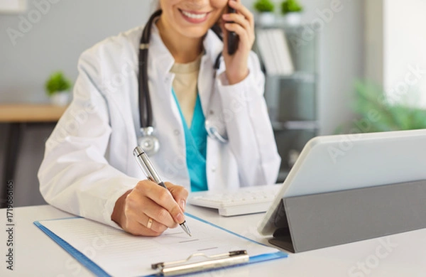Fototapeta Doctor, nurse, medical professional at desk in office writing on clipboard, talking mobile phone, in white lab coat, stethoscope, focused work and multitasking, managing tasks and communication, call