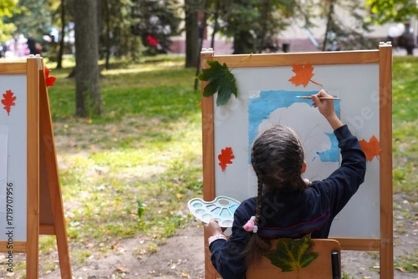 Obraz Child focused on painting at an easel adorned with autumn leaves in a park. The artwork process is underway, showcasing creativity and concentration amidst nature's colorful backdrop.