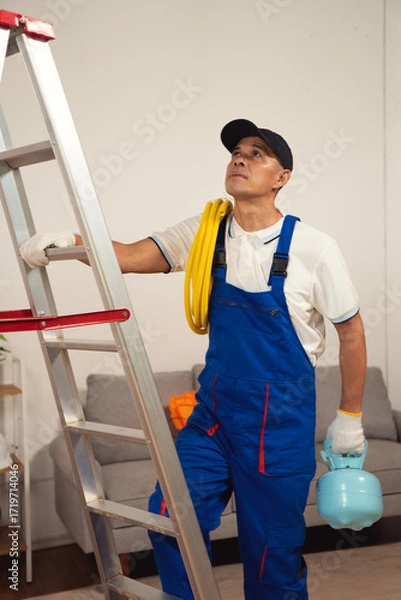 Fototapeta Handyman's Ascent: A skilled handyman in professional attire ascends a ladder indoors, carrying a water sprayer and a cable, ready to tackle maintenance. This image depicts readiness.
