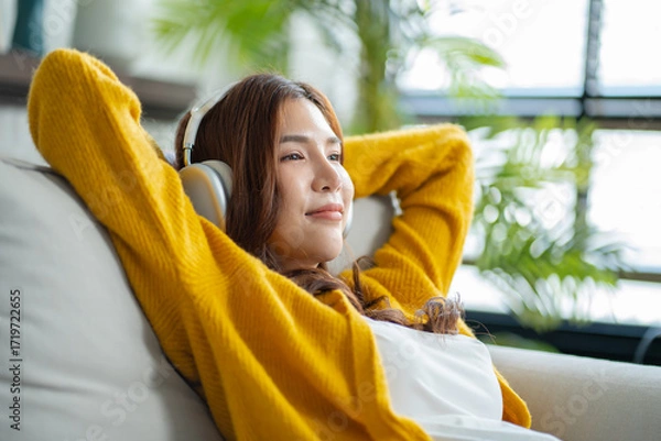 Fototapeta Happy young asian woman use headphones and relaxing at home.Female smile sitting on sofa,couch and holding mobile smartphone.Listening to music on lazy and radiating joy and relaxation.