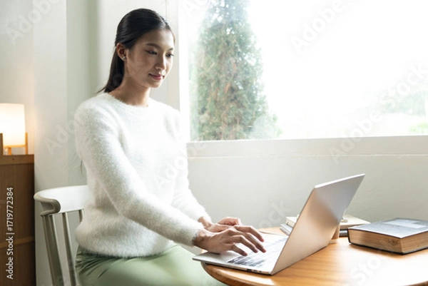 Fototapeta Young Asian woman enjoying a peaceful moment reading a book near window. Calm lifestyle, self-learning, and mindfulness at home.