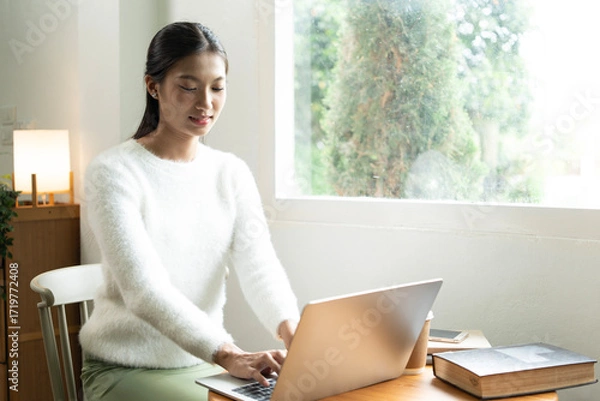 Fototapeta Young Asian woman enjoying a peaceful moment reading a book near window. Calm lifestyle, self-learning, and mindfulness at home.