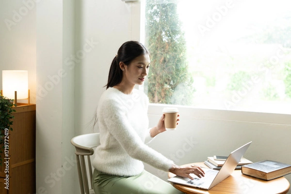 Fototapeta Young Asian woman enjoying a peaceful moment reading a book near window. Calm lifestyle, self-learning, and mindfulness at home.