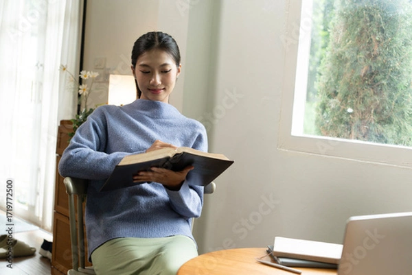 Fototapeta Young Asian woman enjoying a peaceful moment reading a book near window. Calm lifestyle, self-learning, and mindfulness at home.