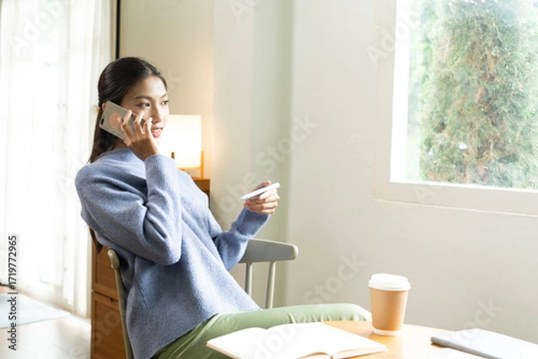 Fototapeta Young Asian woman enjoying a peaceful moment reading a book near window. Calm lifestyle, self-learning, and mindfulness at home.