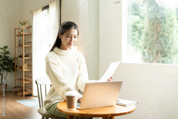 Fototapeta Young Asian woman enjoying a peaceful moment reading a book near window. Calm lifestyle, self-learning, and mindfulness at home.