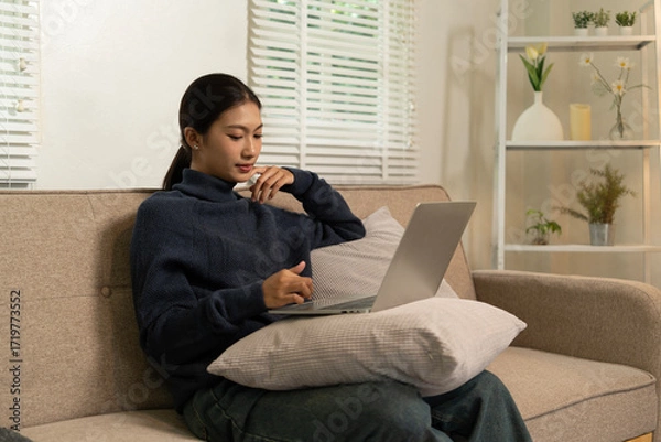 Fototapeta Young Asian woman enjoying a peaceful moment reading a book near window. Calm lifestyle, self-learning, and mindfulness at home.