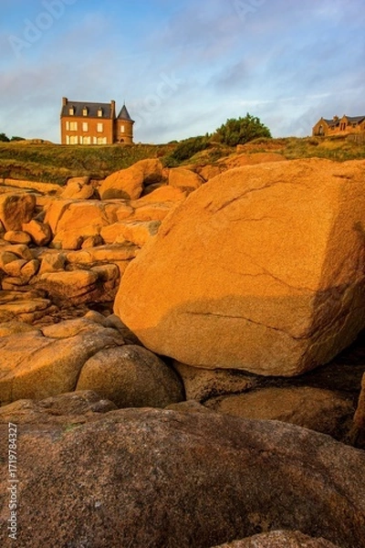 Fototapeta Granite rocks glowing in warm sunset light along the coast of Brittany, France. A historic seaside house with a turret stands in the background under a peaceful evening sky.