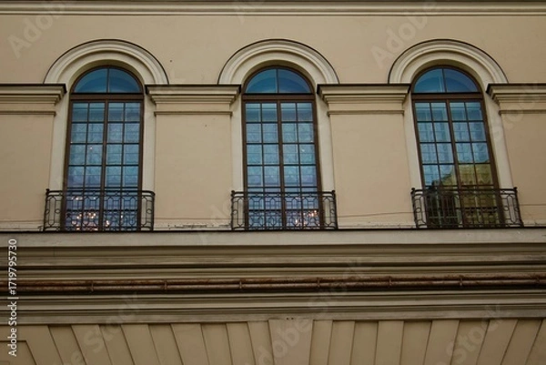 Fototapeta Three tall arched windows of a historic building with wrought iron balcony railings, reflecting the sky and city lights, emphasizing classical architecture elegance.