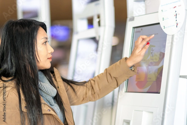 Fototapeta A young woman using self check-in kiosks in airport terminal.