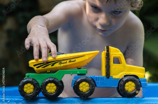 Fototapeta A boy plays with a motorboat and loads it onto a truck on the edge of a plastic swimming pool in a summer garden