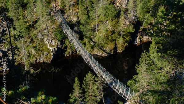 Fototapeta A breathtaking view of the Canyon Lake Julma Olkky in Hossa National Park, Finland, showcasing a wooden bridge crossing a serene body of water surrounded by lush greenery.