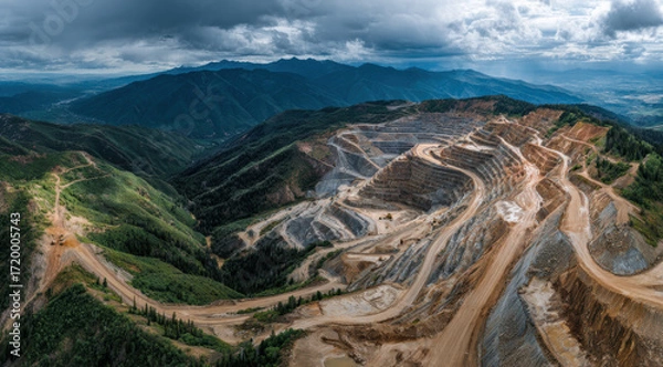 Fototapeta Aerial view of large open pit mine with terraced levels, surrounded by lush green mountains under cloudy sky