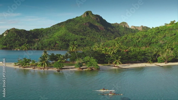 Obraz Two people paddling an outrigger canoe near tropical island with white sand beach, green towering mountains in background. Active sports lifestyle. French Polynesia. remote wild nature paradise aerial