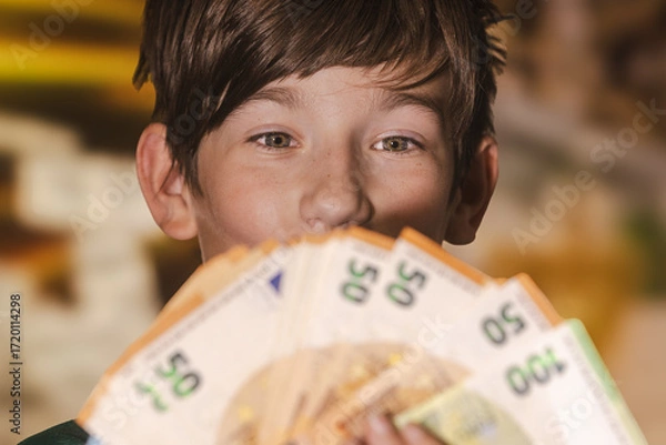Fototapeta Smiling boy proudly holding a fan of euro banknotes in 20, 50, and 100 denominations, symbolizing wealth, savings, finance, and prosperity.