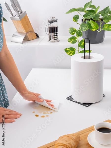 Fototapeta A person's hand uses a paper towel to clean a coffee spill from a white kitchen table, with a paper towel holder and coffee utensils visible in the background.