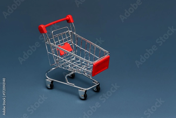 Fototapeta Shortage, crisis. Hunger. Empty metal grocery shopping cart with red handles and inserts on a solid blue background. Horizontal photo