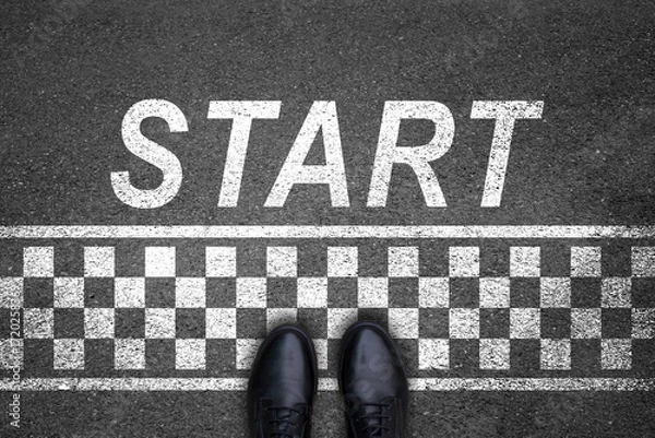 Fototapeta A pair of feet on a tarmac road with white print of the word start for the concept of starting point and the finish line.