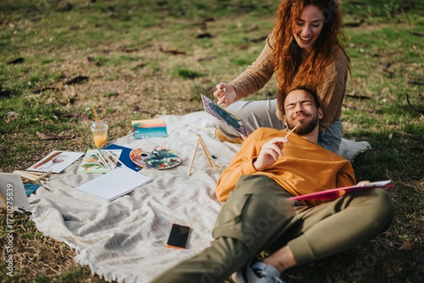 Fototapeta Cheerful couple outdoors enjoying a sunny day by painting together on a blanket surrounded by art supplies. The atmosphere radiates creativity, relaxation, and togetherness in a nature-filled setting.