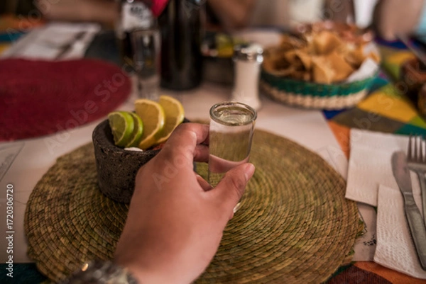 Fototapeta POV photo with hand and delicious crystalline mezcal in its glass caliber, accompanied by worm salt, oranges, and lemon served in a small molcajete.
