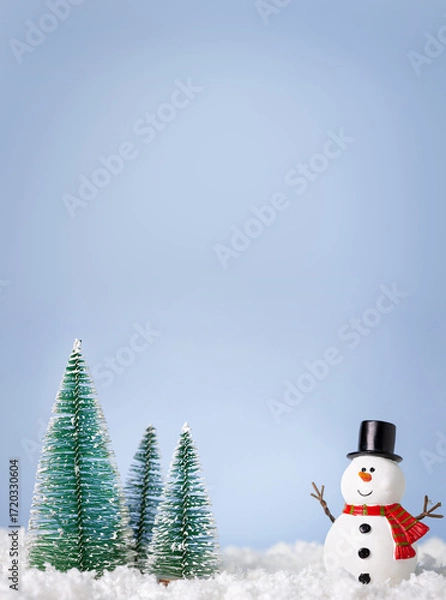 Fototapeta Christmas greeting card. Snowman and new year trees on snow over blue paper background with copy space