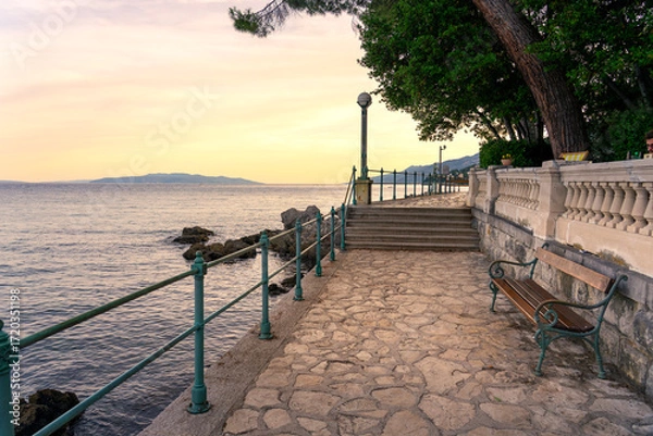 Fototapeta beautiful seaside promenade at sunset with stone pavement, benches, railing, and a calm sea. This is the Lungomare promenade in Opatija, Croatia