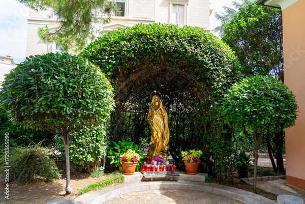 Fototapeta golden statue of the Virgin Mary Madonna placed under a green leafy arch with potted plants and red devotional candles in front. religious shrine or grotto in an outdoor garden in Opatija Croatia