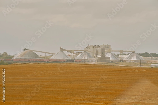 Fototapeta A potash plant with multiple dome-shaped storage buildings and conveyors viewed across farmland under cloudy skies in Saskatchewan, Canada.
