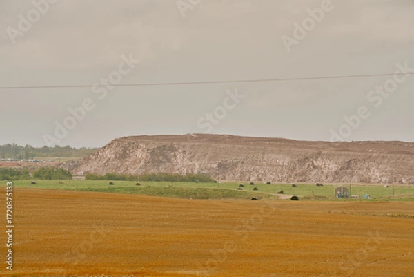Fototapeta A large red potash tailings pile stretches across the horizon behind farmland with hay bales and a small building under cloudy skies in Saskatchewan, Canada.