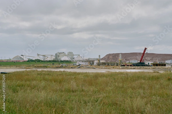 Fototapeta A potash plant with dome storage buildings, conveyor systems, and a red crane beside a wetland and tailings pile under cloudy skies in Saskatchewan, Canada.