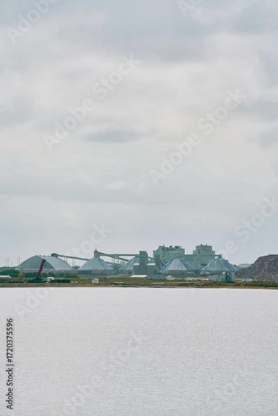 Fototapeta Industrial potash facility with dome storage buildings and conveyor systems viewed across a body of water in Saskatchewan, Canada, under a dramatic cloudy sky.