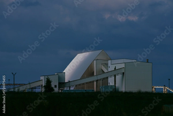 Fototapeta Main processing building of a potash plant in Saskatchewan, Canada, with white industrial architecture, ventilation stacks, and smoke rising under dramatic dark clouds.
