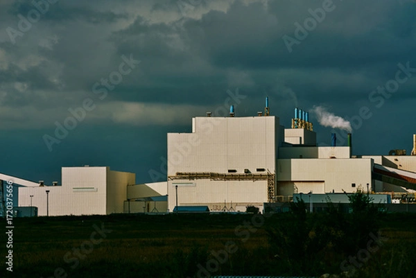 Fototapeta Main processing building of a potash plant in Saskatchewan, Canada, with white industrial architecture, ventilation stacks, and smoke rising under dramatic dark clouds.