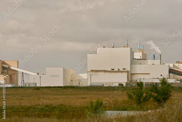 Fototapeta Main processing building of a potash plant in Saskatchewan, Canada, with white industrial architecture, ventilation stacks, and smoke rising under dramatic dark clouds.