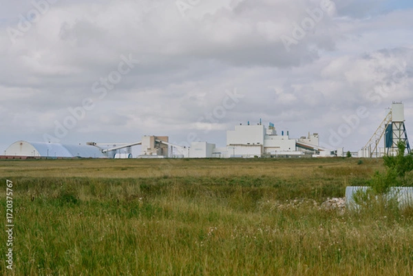 Fototapeta Main processing building of a potash plant in Saskatchewan, Canada, with white industrial architecture, ventilation stacks, and smoke rising under dramatic dark clouds.