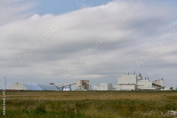Fototapeta Modern industrial mining facility in Saskatchewan, Canada, featuring white factory buildings, conveyor systems, and a tall shaft tower against a cloudy sky with grassy foreground.