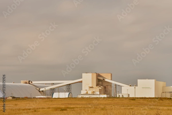 Fototapeta Modern industrial mining facility in Saskatchewan, Canada, featuring white factory buildings, conveyor systems, and a tall shaft tower against a cloudy sky with grassy foreground.