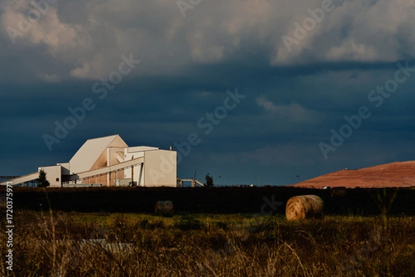 Fototapeta Industrial potash facility in Saskatchewan, Canada, with white processing buildings, conveyor systems, hay bales in the foreground, and tailings pile under dramatic cloudy sky.