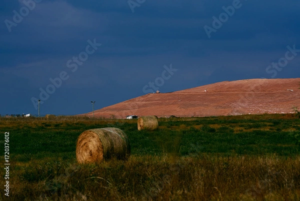 Fototapeta Round hay bales on farmland in Saskatchewan, Canada, with a large potash tailings pile in the background under dramatic cloudy sky.